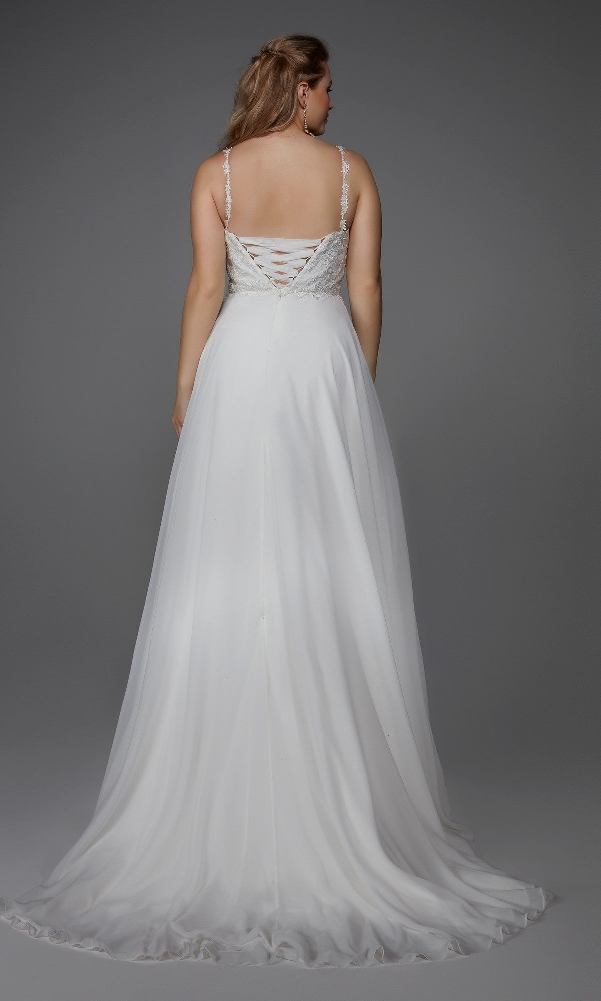 A woman wears the Long White Dress: Alyce 7018, featuring a chiffon a-line skirt and lace-up back, as she stands against a gray background, shown from behind.